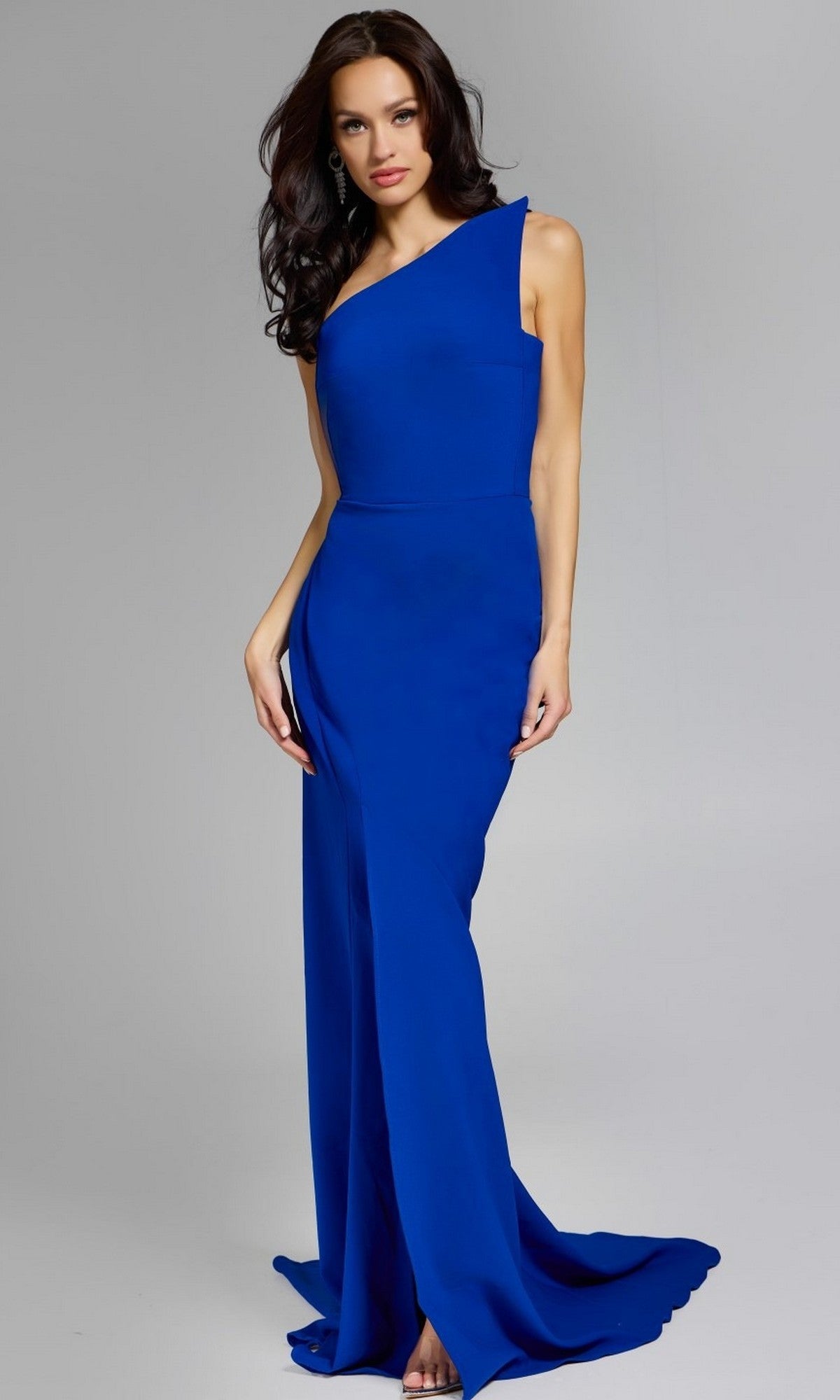 A woman poses in the floor-length, sleeveless Jovani 44001 long prom dress featuring a royal blue one-shoulder design with an asymmetrical neckline, set against a plain gray background.